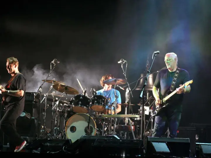 Les membres de Pink Floyd sur scène, David Gilmour à la guitare, Nick Mason à la batterie, et un autre musicien.