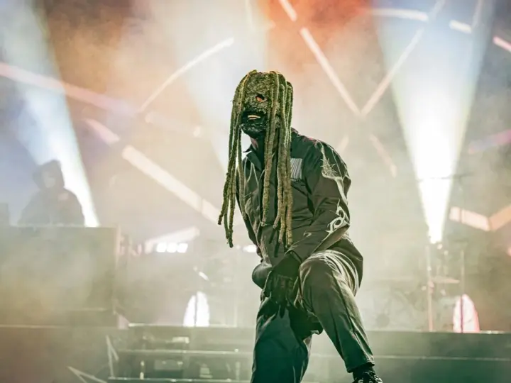 Un membre de Slipknot, masqué et en combinaison, sur scène sous des projecteurs.