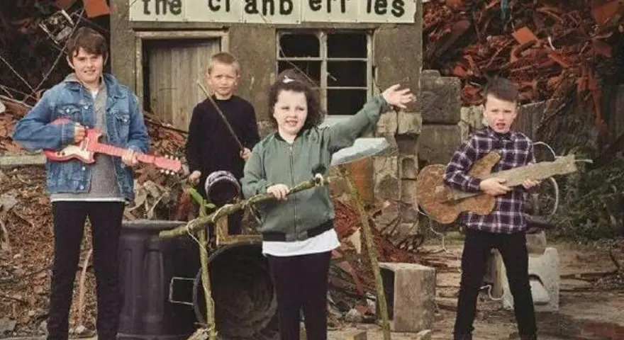 Quatre enfants jouent de la musique devant un panneau "the cranberries". L'un tient une guitare rose, un autre une guitare en bois.