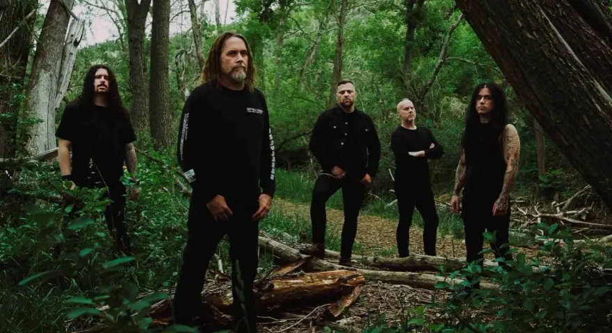 Cinq hommes en noir posent dans une forêt. Le groupe, qui pourrait être Cattle Decapitation, se tient parmi les arbres et la végétation luxuriante.