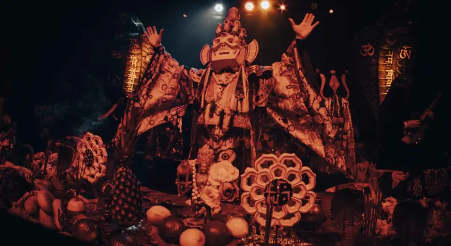 Un personnage masqué, drapé dans des tissus ornés, semble présider un rituel. Autour de lui, des offrandes de fruits, des serpents stylisés et des symboles sacrés évoquent un culte du feu mystérieux.