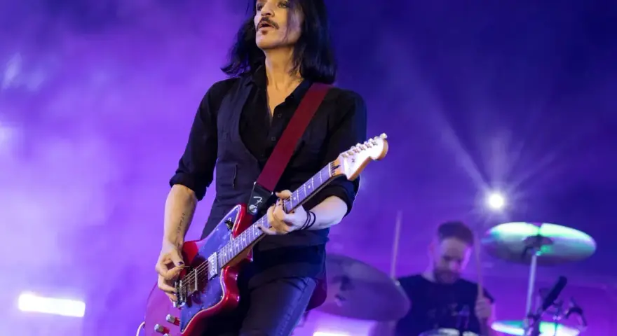 Brian Molko, le chanteur de Placebo, joue de la guitare rouge sur scène, éclairé par des lumières violettes.