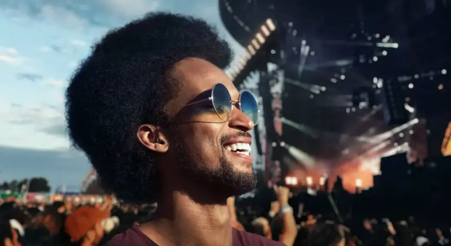 Un homme souriant avec des lunettes de soleil profite d'un concert en plein air. Il porte des bouchons d'oreilles, se demandant pourquoi pas de bouchon concert pour protéger son audition.
