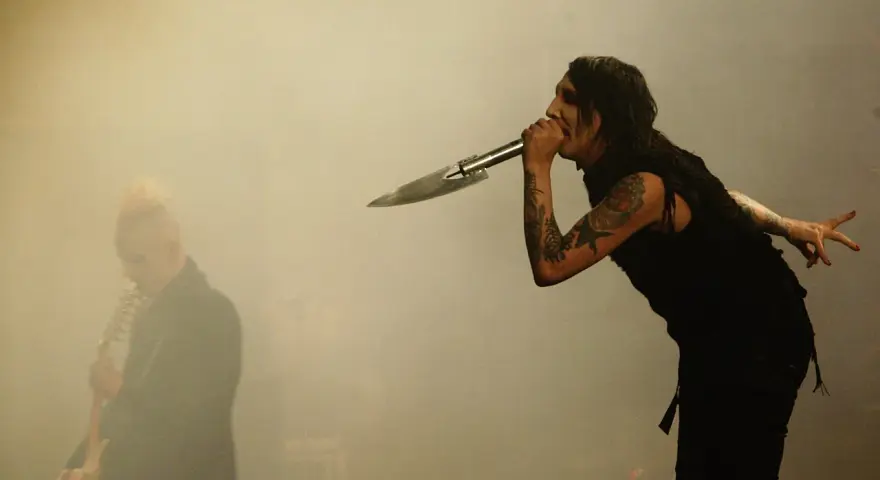 Marilyn Manson chante dans un micro en forme de couteau, un autre musicien est flou à gauche. L'ambiance d'un concert de Marilyn Manson album.