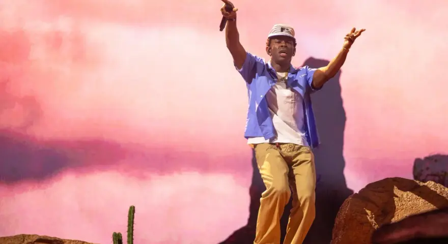 Tyler Okonma, micro en main, danse sur scène devant un décor rose pastel. Il porte une casquette, une chemise bleue ouverte sur un t-shirt blanc et un pantalon beige.