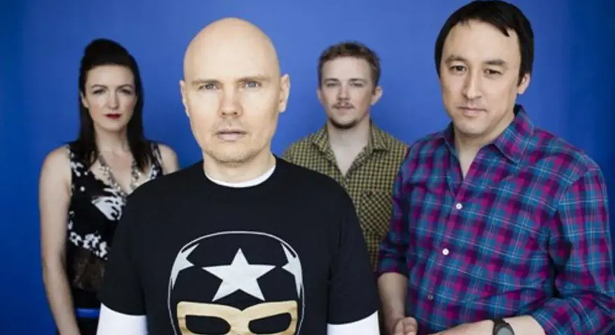 Les membres de The Smashing Pumpkins posent devant un fond bleu. Billy Corgan porte un t-shirt noir avec un masque de lucha.