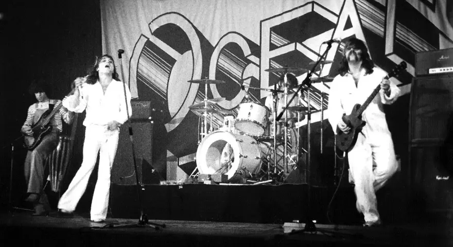 Le groupe Trust en concert, avec le chanteur en blanc au centre, un guitariste à gauche et un autre guitariste à droite. La scène est décorée d'une grande bannière avec le logo du groupe.