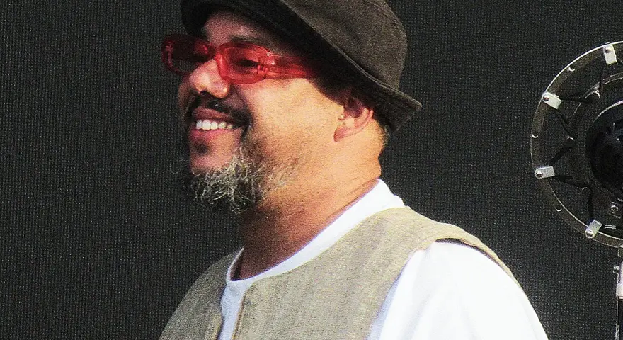 Un chanteur soul souriant, portant un chapeau et des lunettes rouges, se tient devant un microphone.