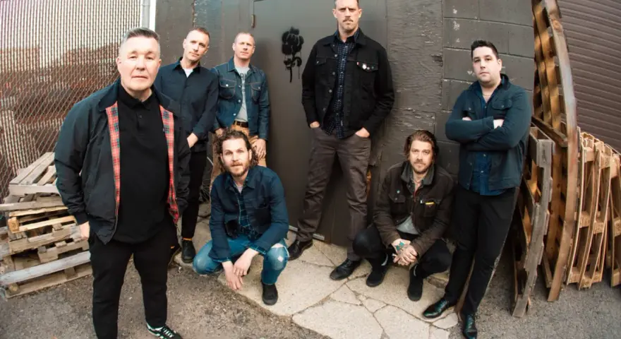 Les membres de Dropkick Murphys posent devant un mur sombre, certains accroupis, d'autres debout, prêts pour leur prochain concert.