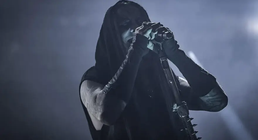 Chanteur de Dimmu Borgir, visage encapuchonné et maquillé, chantant dans un micro, ambiance sombre et enfumée.