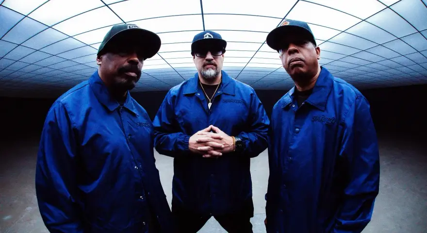 Les membres de Cypress Hill, B-Real, Sen Dog et DJ Muggs, posent ensemble, portant des vestes bleues assorties sous un plafond géométrique.