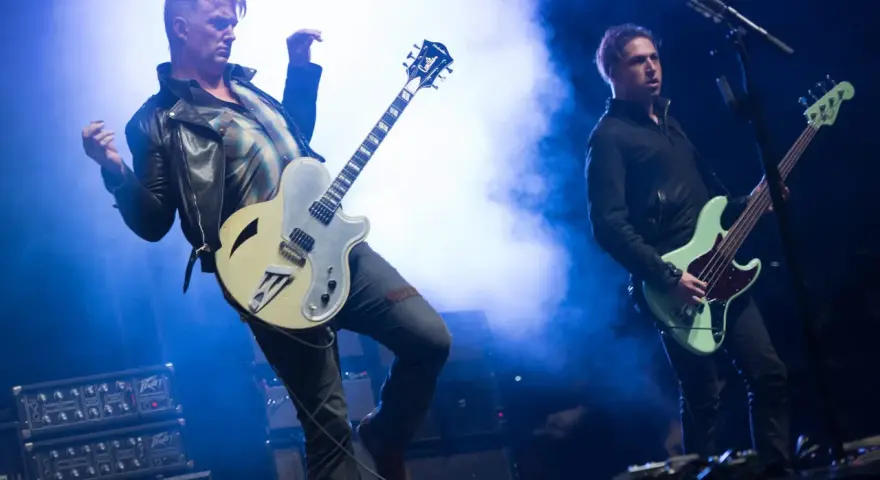Deux membres de Queens of the Stone Age en pleine performance, l'un jouant de la guitare, l'autre de la basse, sous des éclairages bleus intenses.