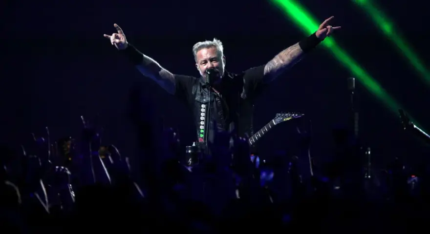 James Hetfield, un des membres de Metallica, salue la foule avec les cornes du diable, éclairé par des faisceaux verts.