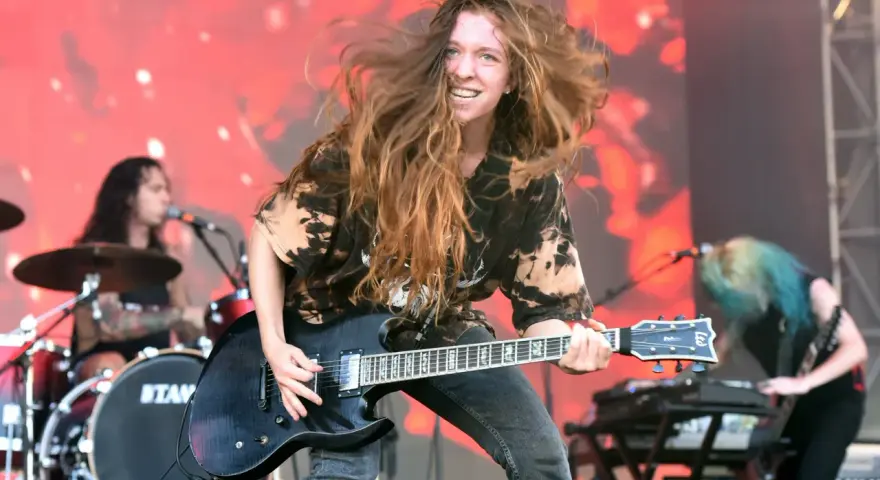 Reba Meyers, guitariste au look sauvage, fait vibrer sa guitare électrique sur scène, cheveux au vent, devant un fond rougeoyant.