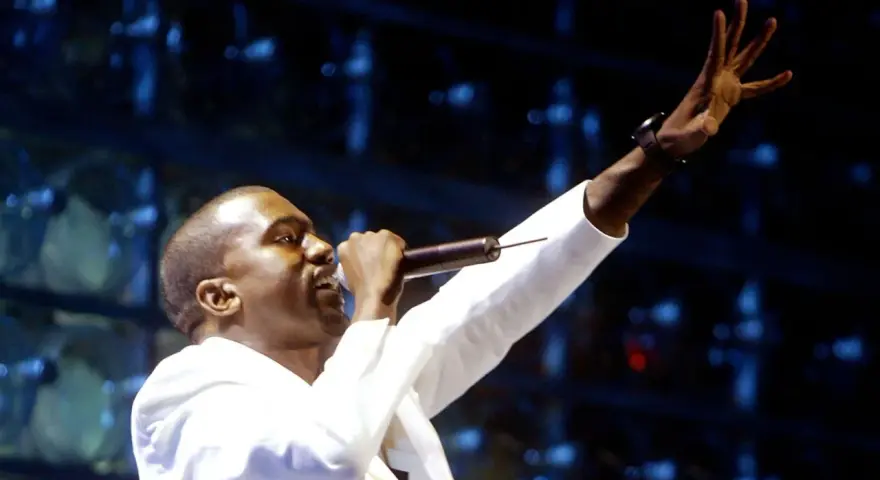 Kanye West, aux origines de sa carrière, chante sur scène, micro à la main, bras levé.