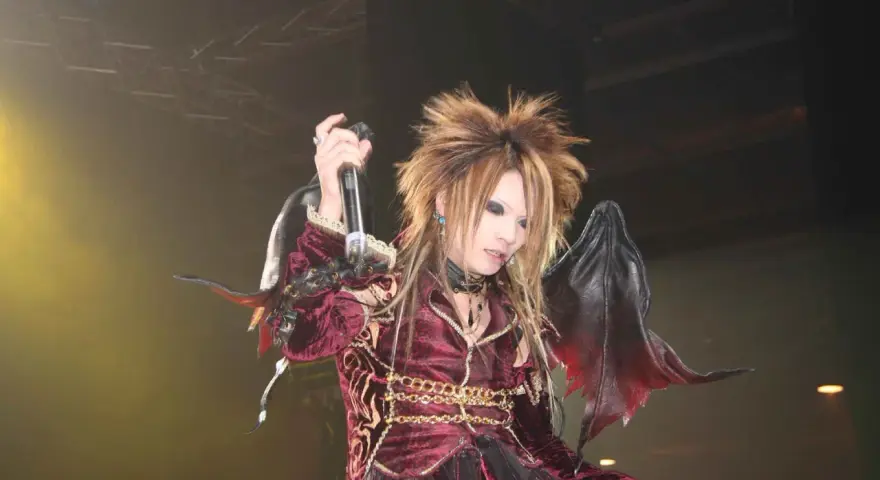 Un membre de X Japan, cheveux ébouriffés, maquillage prononcé, costume sombre et ailes de démon, chante sur scène.