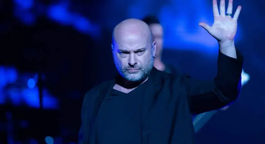 Le chanteur Disturbed, chauve et barbu, lève la main sur scène sous des lumières bleues intenses.