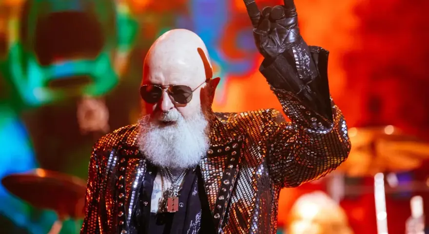 Rob Halford, un des membres de Judas Priest, fait le signe des cornes sur scène, vêtu d'une veste scintillante.