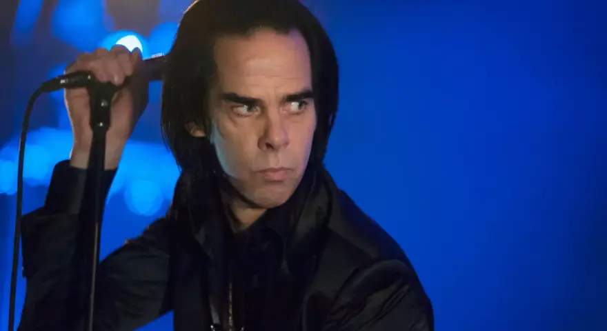Nick Cave, chanteur du groupe australien des années 80, sur scène, micro à la main, regard intense sur fond bleu.