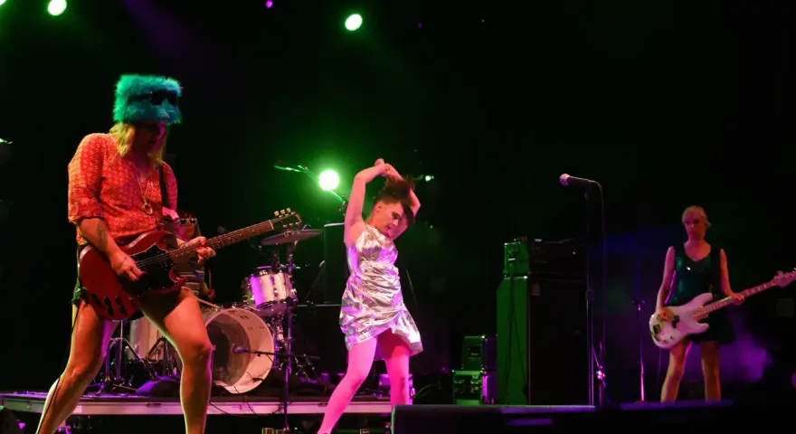 Sur scène, le groupe Bikini Kill enflamme le public. Une guitariste porte un chapeau vert fluo, une autre joue de la basse, et une troisième danse avec énergie.