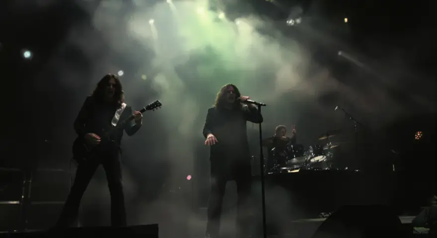 Un groupe de metal sur scène, avec un guitariste à gauche, un chanteur au centre, et un batteur à droite. La scène est enfumée, éclairée par des projecteurs.