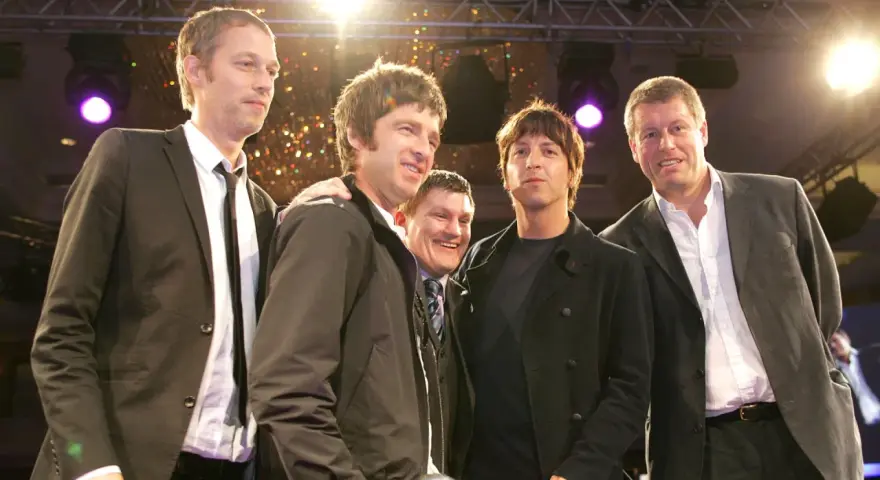 Cinq hommes, dont le groupe Oasis, posent ensemble sur scène, souriants sous les lumières.