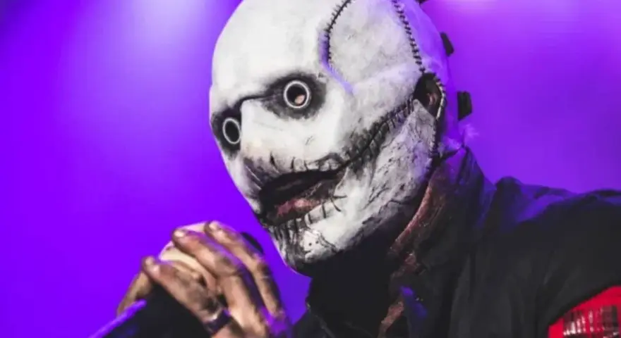 Le chanteur Slipknot, masqué et terrifiant, tient un micro sur scène sous une lumière violette.