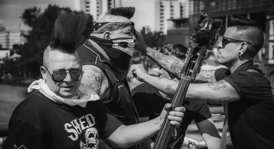 Quatre musiciens au look punk, dont un avec un bandana et un masque de crâne, se préparent à jouer. Un d'eux tient une contrebasse, prêt à faire un mad sin.