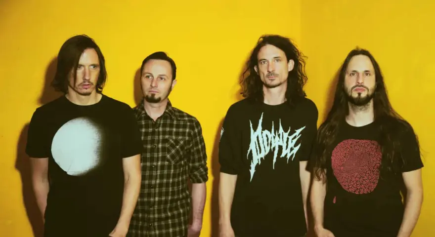 Quatre hommes aux cheveux longs posent devant un fond jaune vif. Leurs t-shirts portent des graphismes distinctifs, évoquant l'énergie brute du magma et la puissance de Gojira.