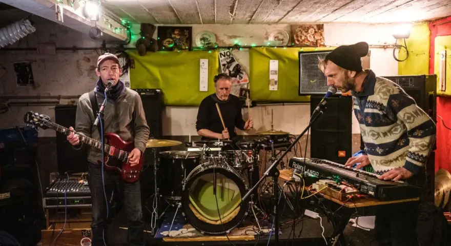 Trois musiciens jouent dans un local sombre, éclairé par des guirlandes. Un guitariste, un batteur et un claviériste, oi boys !