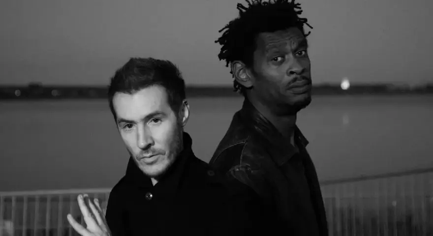 Les membres de Massive Attack, 3D et Daddy G, posent dos à dos, regardant dans des directions opposées, dans une ambiance sombre et contemplative.