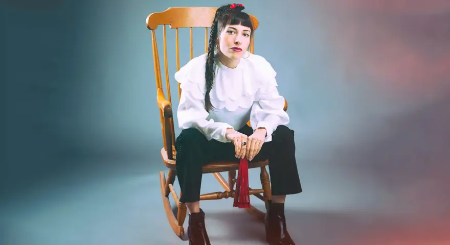 Scout Niblett, assise sur un fauteuil à bascule, porte une blouse blanche à volants et un pantalon noir. Elle tient un pompon rouge.