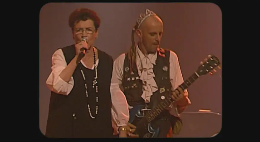 Une chanteuse et un guitariste, peut-être les ramoneurs de menhirs, sur scène. L'homme porte une couronne et une tenue excentrique.