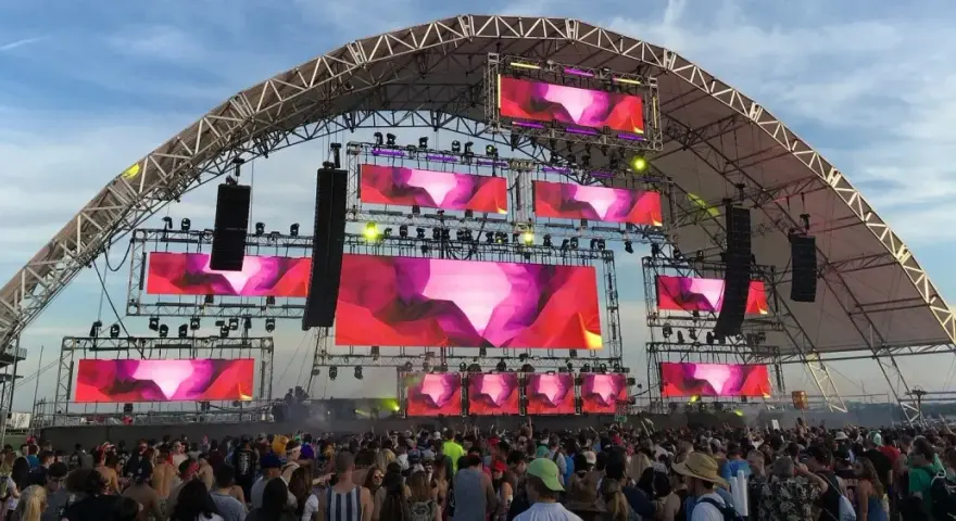 Scène de festival avec écrans LED diffusant des visuels abstraits roses et violets. Une foule danse sous la structure, attendant leur artiste deep house préféré.