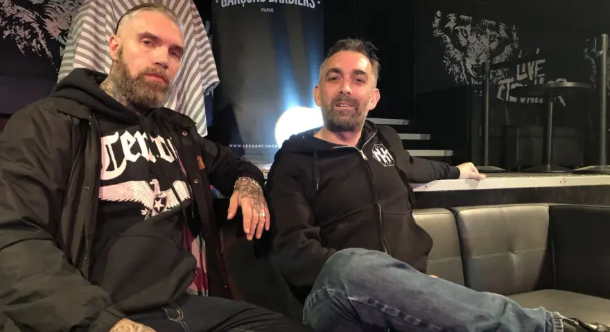 Deux hommes, dont un possible chanteur de Mass Hysteria, posent sur un canapé. L'un porte un sweat à capuche noir avec un aigle.