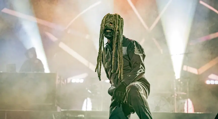 Un membre de Slipknot, masqué et en combinaison, sur scène sous des projecteurs.