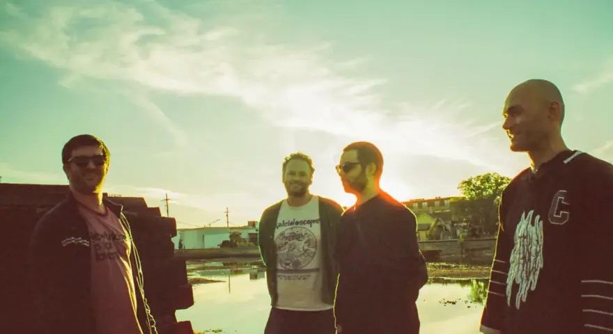 Quatre hommes posent devant un ciel clair, comme un chat pile. L'un porte un t-shirt "kaleidoscope".