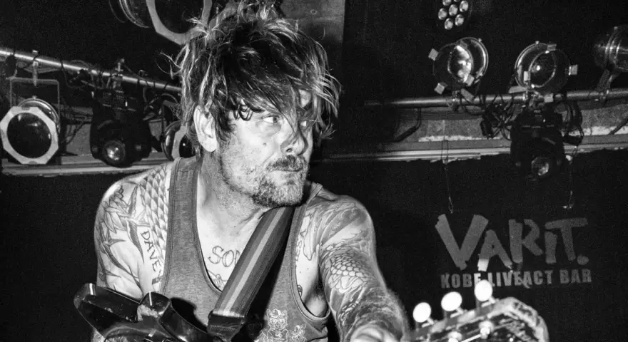 Un guitariste tatoué, cheveux en bataille, sous les projecteurs du VART. Kobe Liveact Bar.