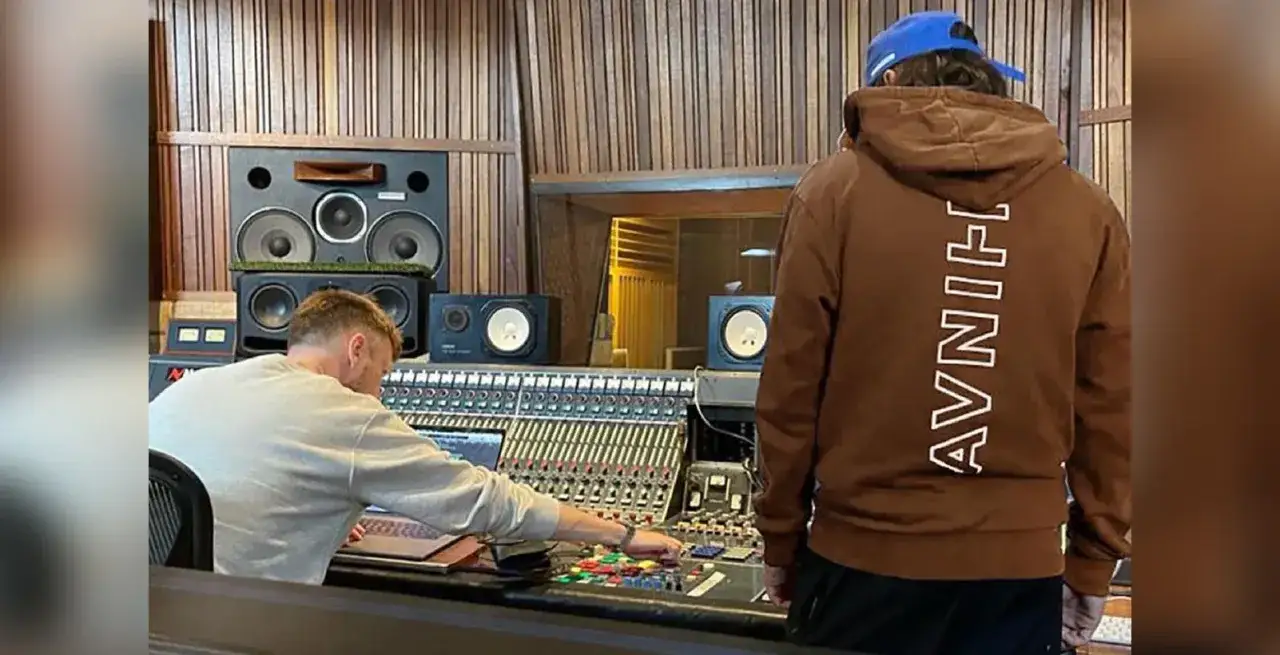 Dans un studio d'enregistrement, un homme ajuste la console de mixage, tandis qu'un autre, portant un sweat marron avec 