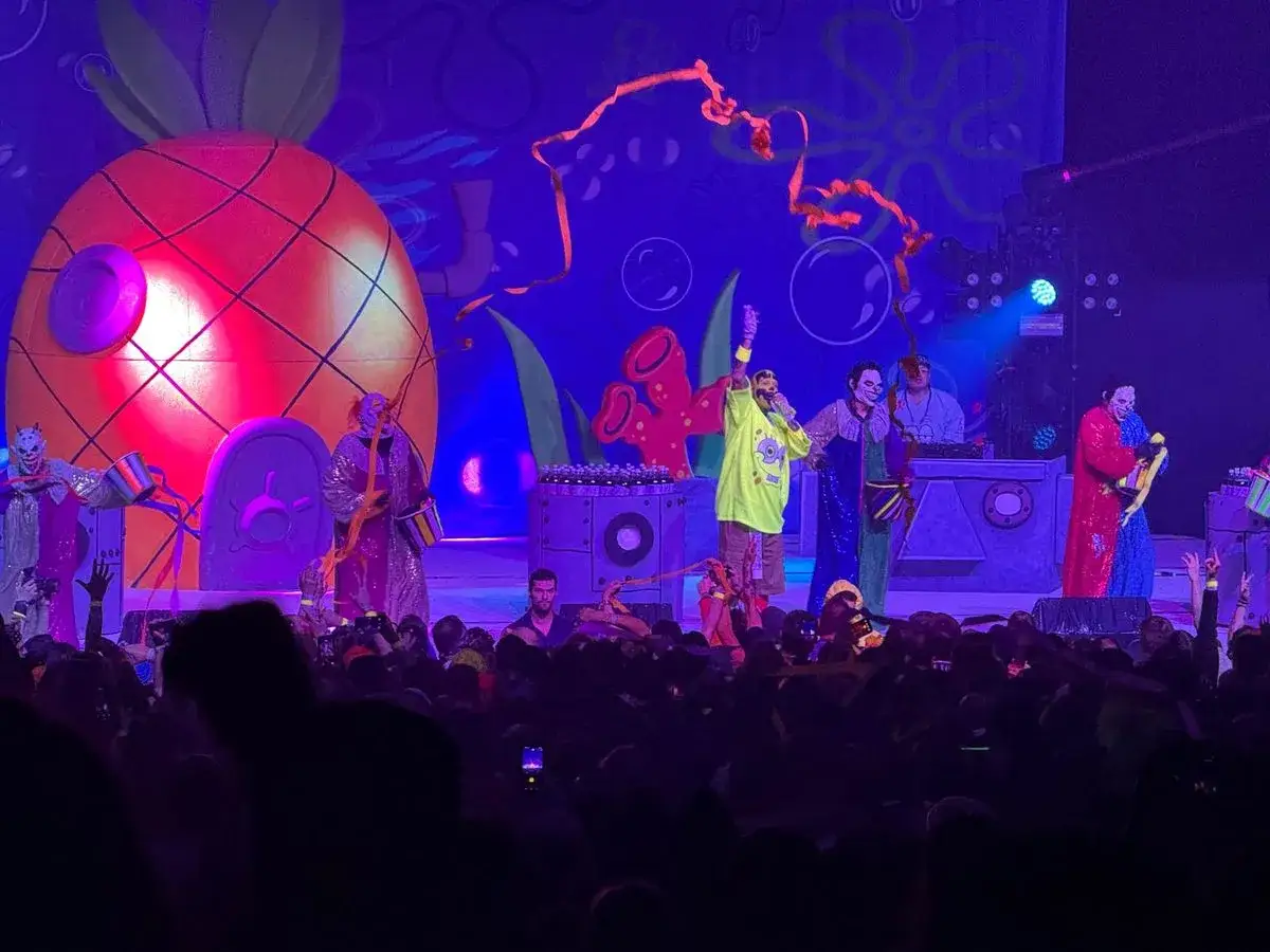 Scène de concert avec un décor de maison d'ananas et des membres de l'Insane Clown Posse en costumes colorés, devant une foule en délire.