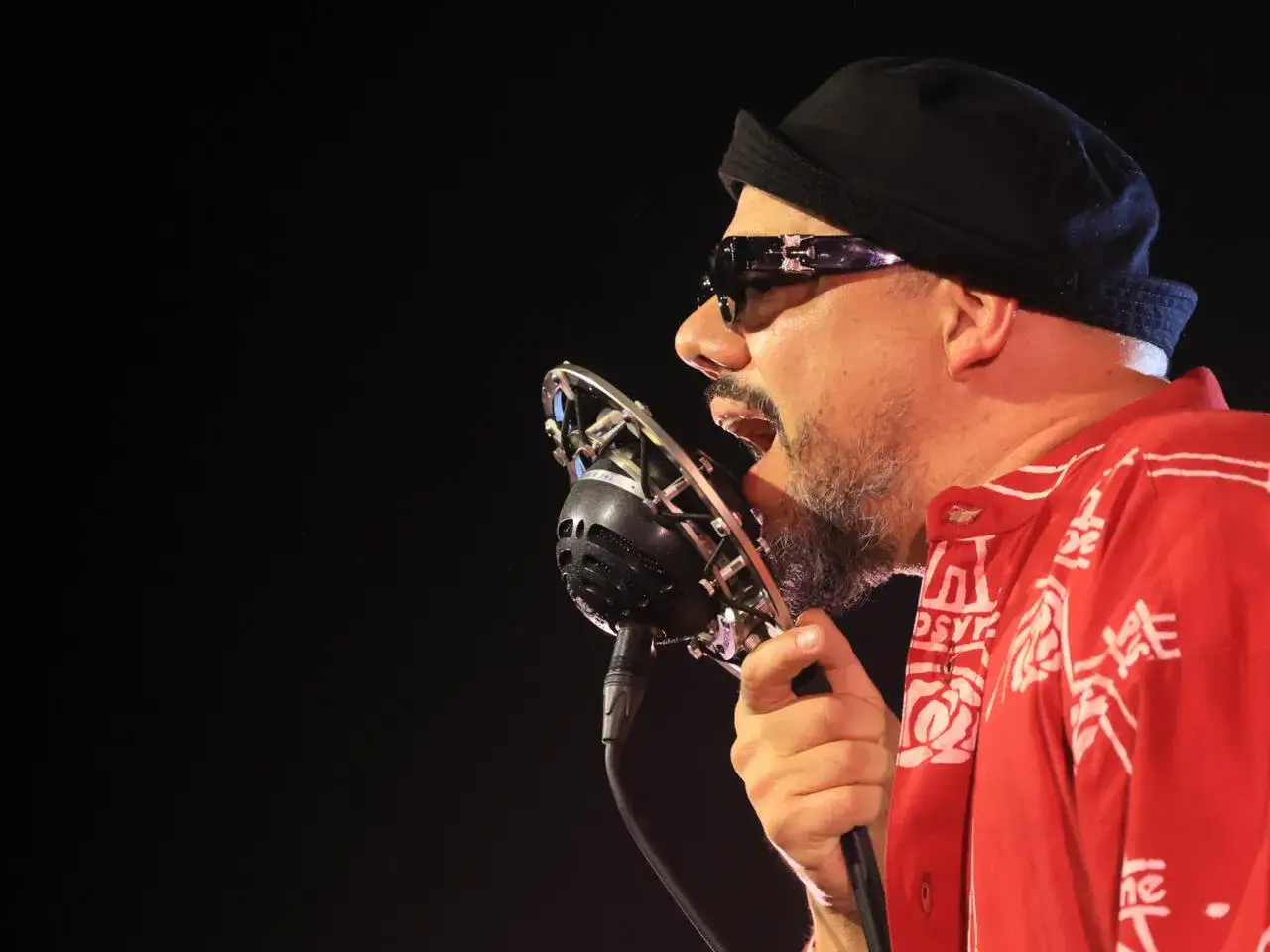 Un chanteur soul, barbe grisonnante, lunettes de soleil et bonnet noir, chante passionn&eacute;ment dans un micro vintage. Sa chemise rouge &agrave; motifs blancs est vibrante.