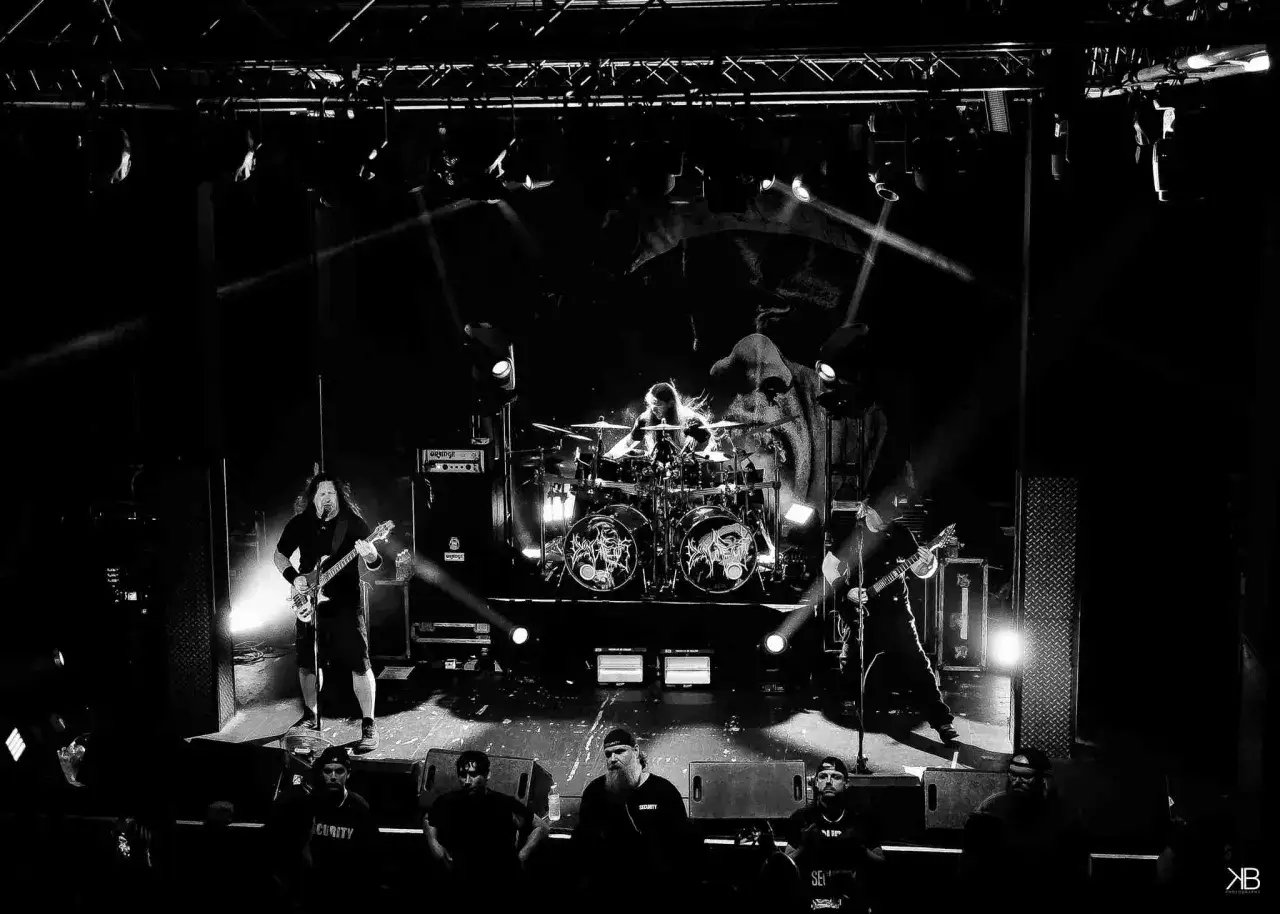 Groupe de metal sur scène, éclairage dramatique. Le batteur est au centre, entouré de symboles sombres rappelant un fœtus mourant.