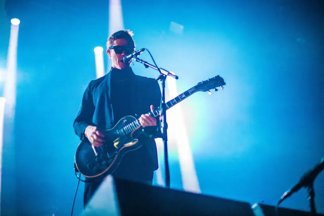 Paul Banks, chanteur du groupe Interpol, joue de la guitare sur sc&egrave;ne, &eacute;clair&eacute; par des projecteurs bleus.