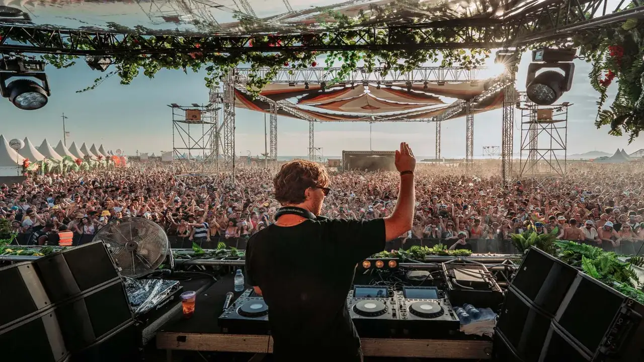 Un DJ, parmi les plus connus, salue une foule immense lors d'un festival en plein air. La scène est décorée de verdure, le soleil brille.