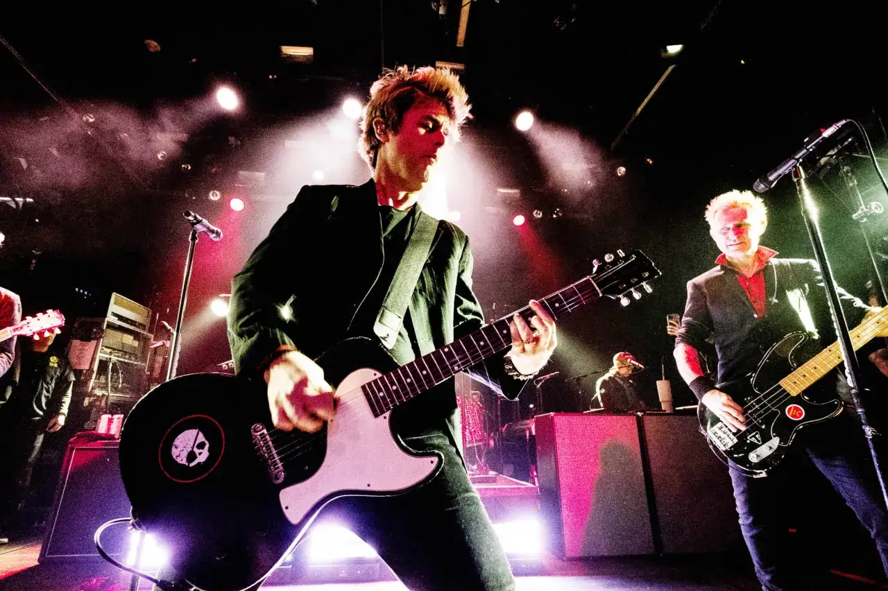 Les membres de Green Day en pleine performance, éclairés par des projecteurs. L'un joue de la guitare, l'autre de la basse.