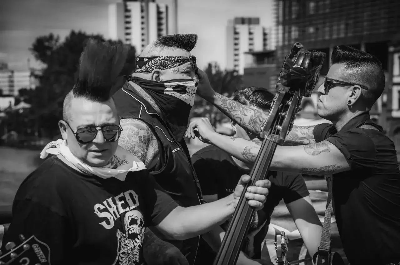 Quatre musiciens au look punk, dont un avec un bandana et un masque de crâne, se préparent à jouer. L'un d'eux tient une contrebasse, prêt à faire un 
