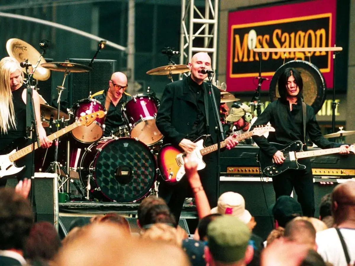 Les membres de The Smashing Pumpkins jouent sur scène. Une guitariste blonde, un batteur chauve, et deux guitaristes chauves et aux cheveux noirs.