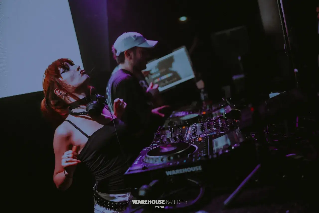 Une dj femme techno, cheveux roux, danse au rythme de la musique, casque sur les épaules, devant une console de mixage.