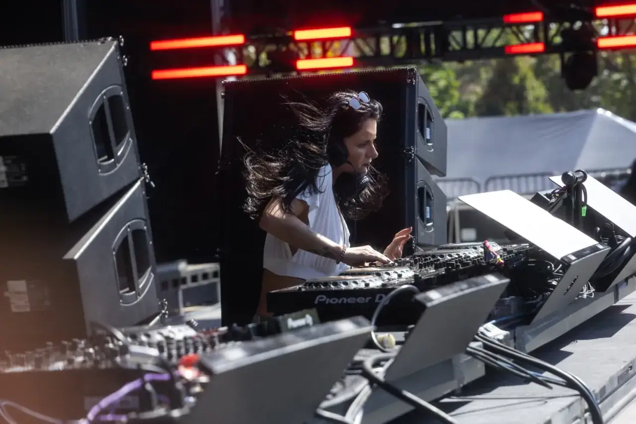 Une dj femme aux cheveux longs et au vent mixe sur scène, entourée de enceintes et de lumières rouges.