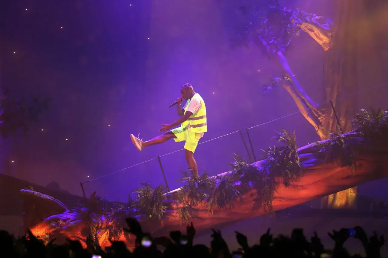 Tyler Okonma, en pleine performance, danse sur une sc&egrave;ne d&eacute;cor&eacute;e d'un arbre g&eacute;ant et de v&eacute;g&eacute;tation luxuriante, sous des lumi&egrave;res violettes.